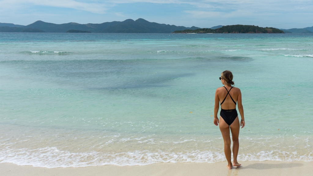A woman standing on Banana Beach in Coron, Philippines and looking out at the bright blue water and mountains.